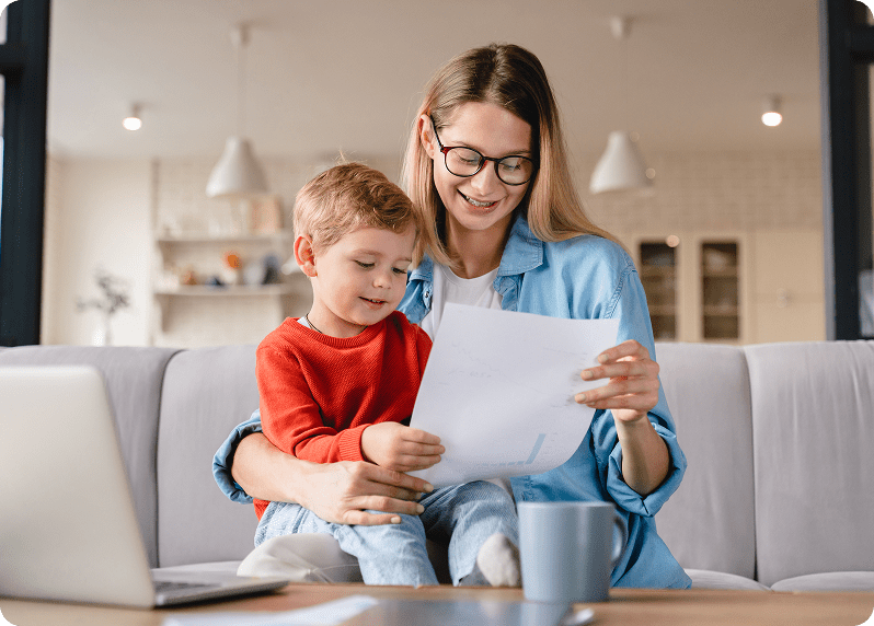 Woman sitting on a couch with a child, both looking at sheets of paper. A laptop and coffee mug are on the table in front of them.
