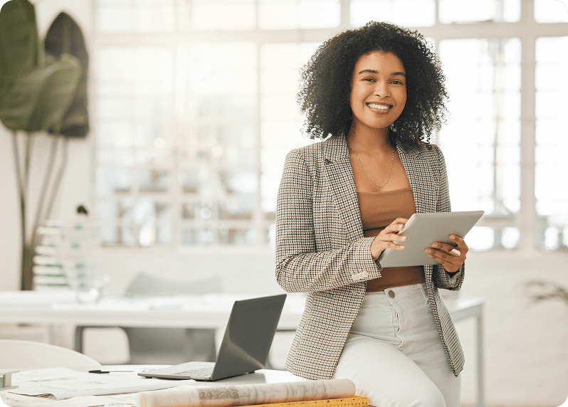Person with curly hair smiling, holding a tablet in one hand, standing in a bright office environment beside an open laptop.