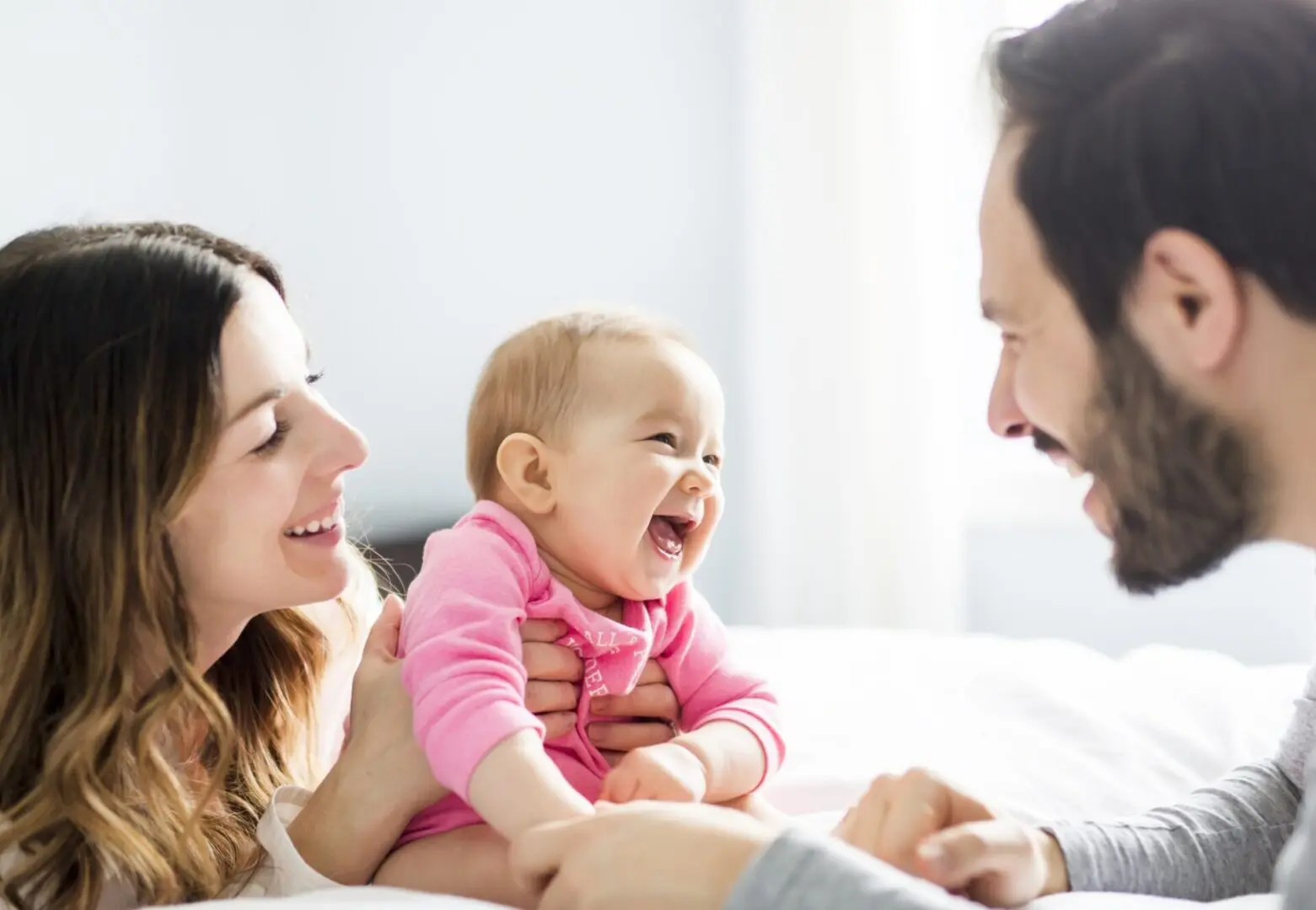 GettyImages-823771718 (1) A couple happily interacts with a laughing baby on a white bed, creating a warm and joyful atmosphere.