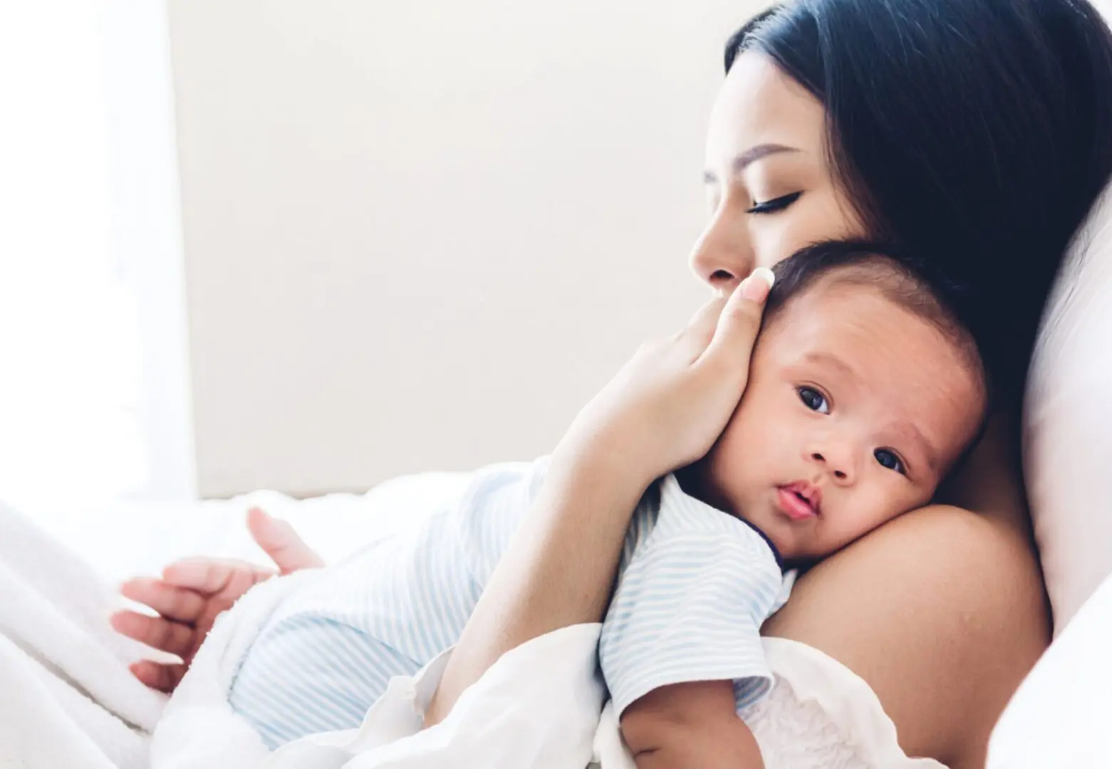 GettyImages-823771718 (2) A woman is lying down, holding a baby close to her chest. The baby is wearing a light blue striped outfit and looking towards the camera. The scene conveys a sense of calm and comfort.