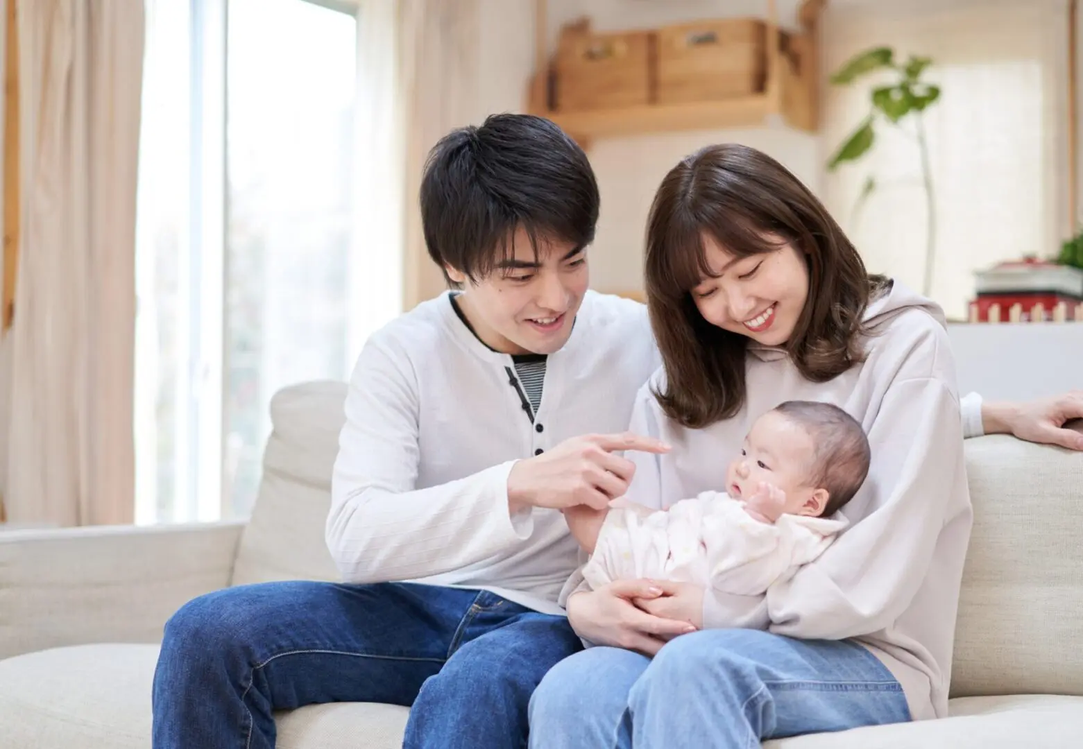 GettyImages-823771718 (3) A couple sits on a couch, smiling at a baby in the woman's arms. The man is pointing at the baby.