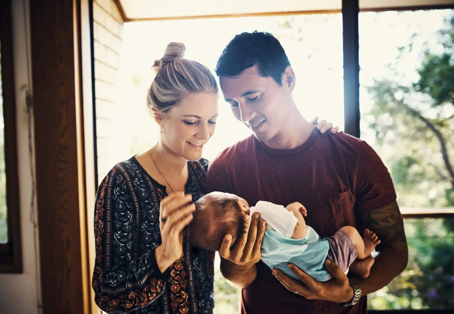 GettyImages-823771718 A couple stands near a window, both smiling as they cradle and look at a baby wrapped in a blanket.