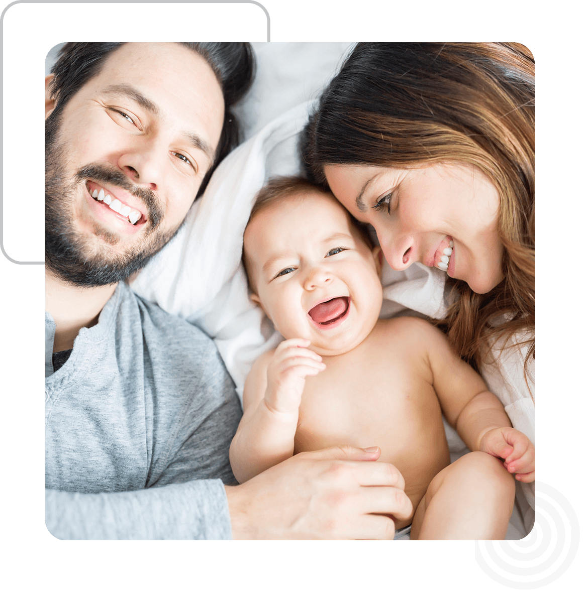 Group 94 (1) A man and woman smile while lying on a bed with a baby between them. The baby is looking at the camera.